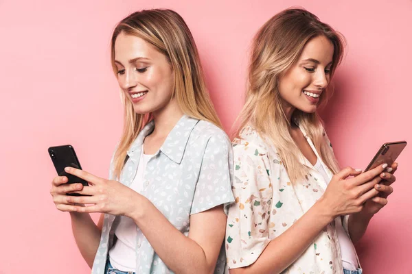 Alegre hermosas rubias mujeres amigas posando aisladas sobre fondo de pared rosa usando teléfonos móviles . — Foto de Stock