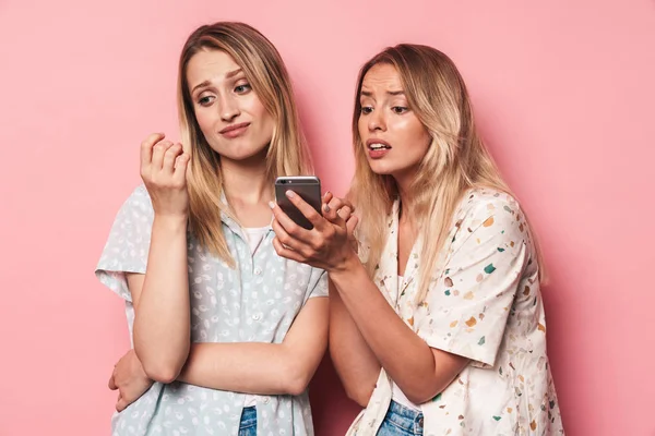ピンクの壁の背景の上に隔離彼女の無関心な女性の友人の携帯電話にディスプレイを示す携帯電話を使用してショックを受けた女の子. — ストック写真