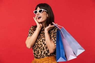 Güzel heyecanlı genç kadın alışveriş çantaları tutan güneş gözlüğü giyen kırmızı arka plan üzerinde izole poz hayvan baskılı gömlek giymiş.