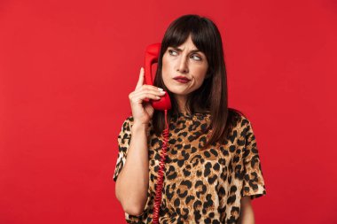 Hayvan baskılı gömlek giymiş düşünen genç kadın telefonla konuşurken kırmızı arka plan üzerinde izole poz.
