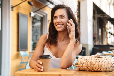 Rahat kafe açık havada otururken kağıt fincan kahve içme ve cep telefonu konuşurken güzel memnun kadın Portresi