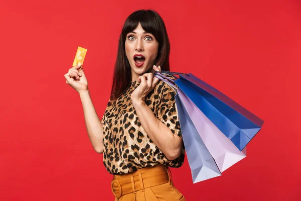 Chockad ung kvinna klädd i djur tryckt skjorta poserar isolerade över röd bakgrund hålla kreditkort och påsar shopping. — Stockfoto