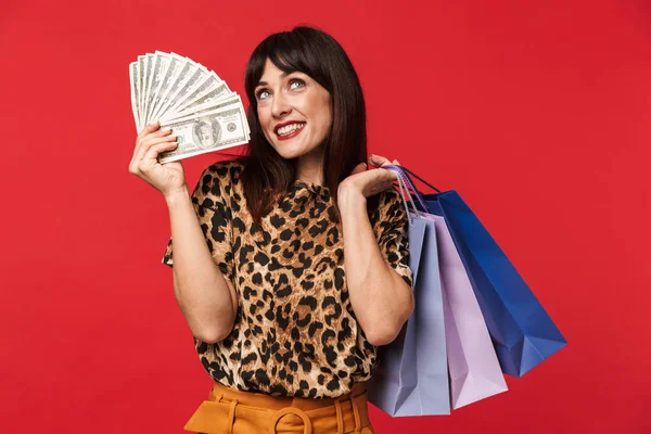 Красива щаслива молода жінка, одягнена у друковану футболку з тваринами, позує ізольовано на червоному тлі, тримаючи гроші та сумки для покупок . — стокове фото