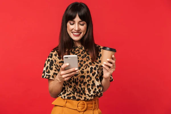 Hermosa joven feliz vestida con camisa animal impresa posando aislada sobre fondo rojo bebiendo café usando teléfono móvil . — Foto de Stock