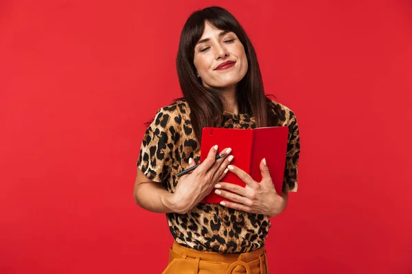 Mooie jonge vrouw gekleed in dierlijke gedrukte shirt poseren geïsoleerd over rode achtergrond schrijven notities in notebook. — Stockfoto