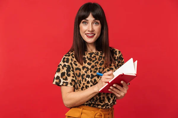 Mooie jonge vrouw gekleed in dierlijke gedrukte shirt poseren geïsoleerd over rode achtergrond schrijven notities in notebook. — Stockfoto