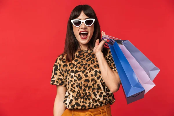 Krásná vzrušená mladá žena oblečená v tištěném zvířecího tričku, která na červeném pozadí nosí sluneční brýle držící nákupní tašky. — Stock fotografie