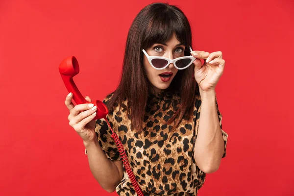 Mooie jonge vrouw gekleed in dierlijke gedrukte shirt poseren geïsoleerd over rode achtergrond dragen zonnebril praten telefonisch. — Stockfoto