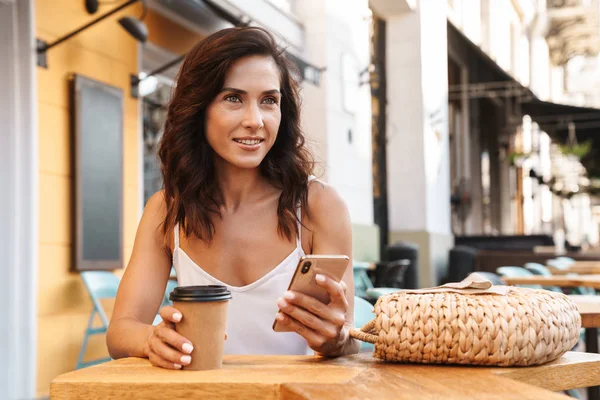 Portré vonzó barna nő szalmakazsák iszik kávét papírpohár és használata cellphone ülve hangulatos kávézóban — Stock Fotó