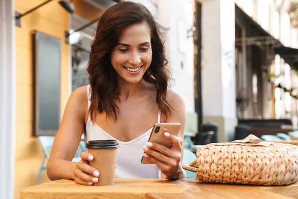 Portré szép elégedett nő iszik kávét papír poharat és használata cellphone ülve hangulatos kávézóban a szabadban — Stock Fotó