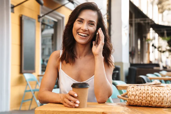 Portret van gelukkig tevreden vrouw drinken van koffie uit de papieren beker en praten op mobiele telefoon terwijl zittend in gezellig café buitenshuis — Stockfoto