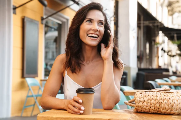 Rahat kafe açık havada otururken kağıt fincan kahve içme ve cep telefonu konuşurken mutlu güzel kadın Portresi