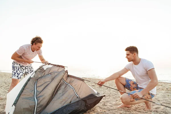 Due uomini felici che costruiscono una tenda sulla spiaggia — Foto Stock