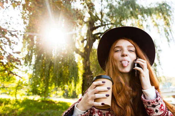 コーヒーを飲んで携帯電話で話す秋の公園で陽気な陽気な若い学生赤毛の女の子を喜んで喜んで. — ストック写真