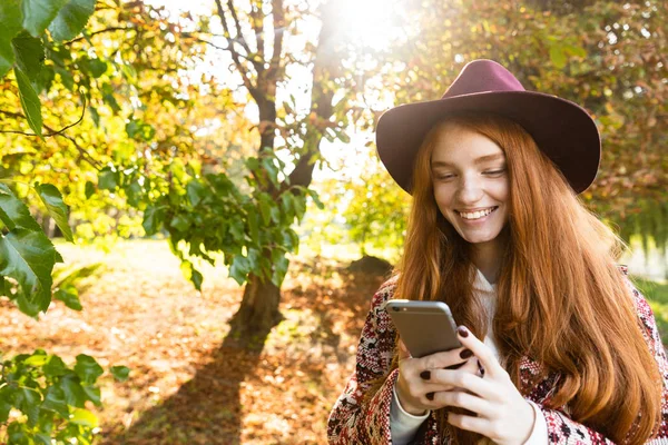 携帯電話を使って秋の公園で笑顔キューティー若い学生赤毛の女の子. — ストック写真
