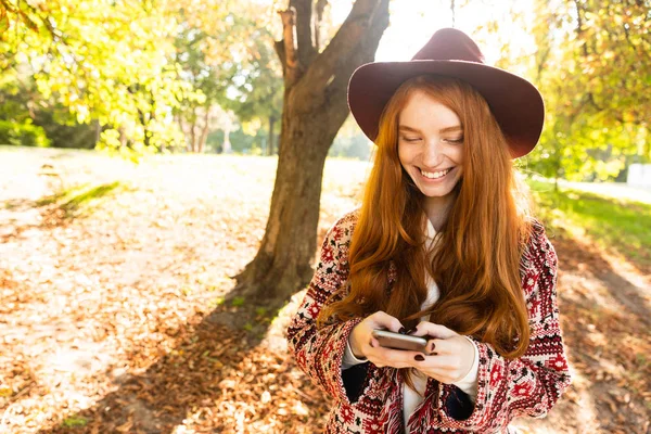 Sonriente chica linda joven estudiante pelirroja en el parque de otoño utilizando el teléfono móvil . —  Fotos de Stock