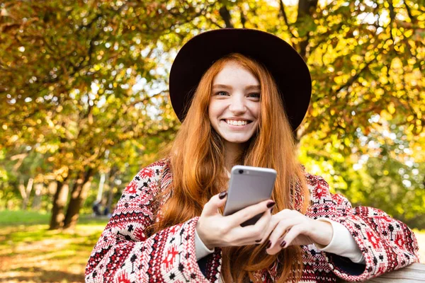 Sonriendo alegre joven estudiante pelirroja en el parque de otoño utilizando el teléfono móvil sentarse en el banco . — Foto de Stock