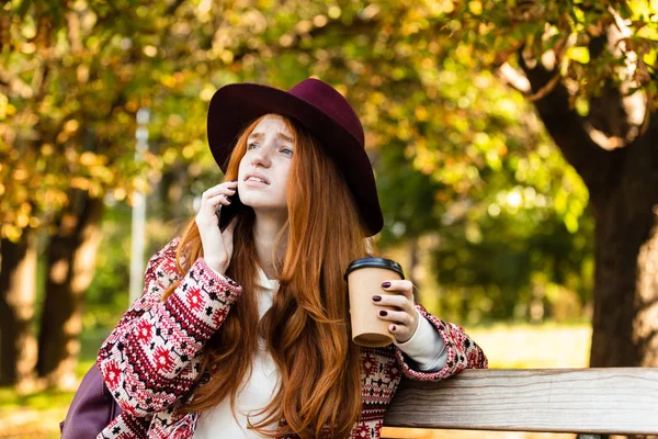 Triste joven estudiante pelirroja chica en otoño parque hablando por teléfono móvil beber café . — Foto de Stock
