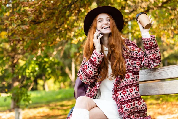 Pozytywny zadowolony szczęśliwy wesoły młody student rudowłosy dziewczyna w Autumn Park rozmowa przez zdejmowany telefon picie Kawa. — Zdjęcie stockowe