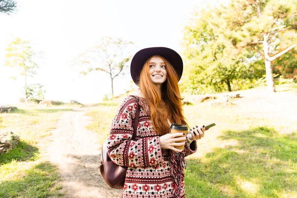 Ładny pozytywny młody student rudowłosy dziewczyna w Autumn Park za pomocą zdejmowany telefon. — Zdjęcie stockowe