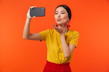 Turuncu duvar arka plan üzerinde cep telefonu ile konuşurken izole poz mutlu genç kadın selfie çekmek.