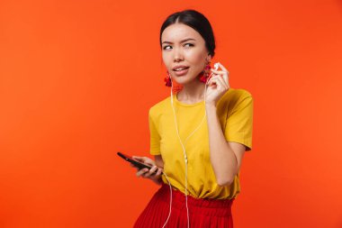 Güzel ciddi karışık genç kadın cep telefonu dinleme müzik kullanarak turuncu duvar arka plan üzerinde izole poz.