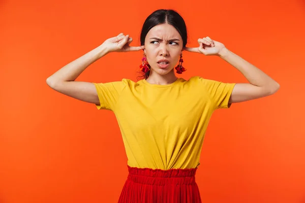 Mooie displease jonge vrouw poseren geïsoleerd over oranje muur achtergrond dekking oren. — Stockfoto