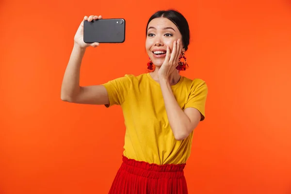 Счастливая молодая женщина позирует изолированно на фоне оранжевой стены разговаривая по мобильному телефону сделать селфи . — стоковое фото