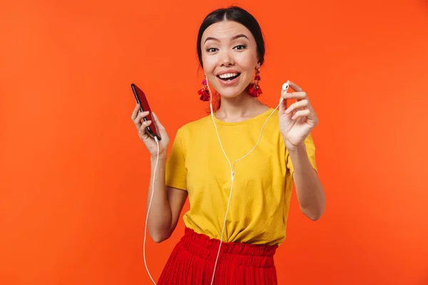 Hermosa joven posando aislada sobre fondo de pared naranja utilizando el teléfono móvil escuchando música . —  Fotos de Stock