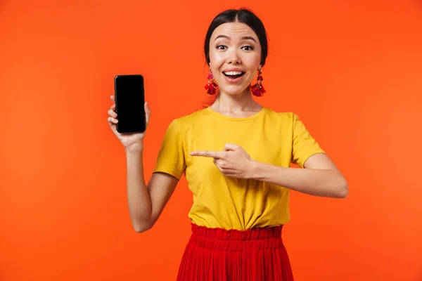 Izgatottan boldog fiatal nő pózol elszigetelt narancssárga fal háttérben mutatja megjelenítését mobiltelefon. — Stock Fotó