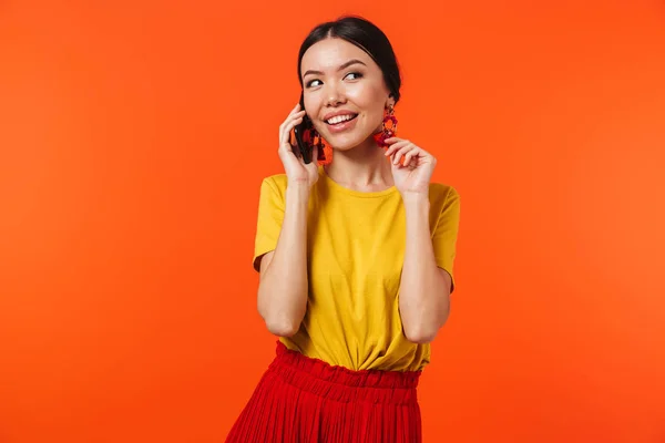 Krásná šťastná mladá žena, která pózovala na pozadí oranžové zdi mluvící mobilním telefonem. — Stock fotografie