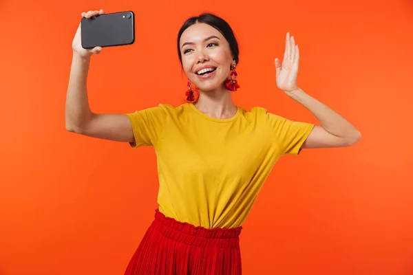 Счастливая молодая женщина позирует изолированно на фоне оранжевой стены разговаривая по мобильному телефону сделать селфи . — стоковое фото