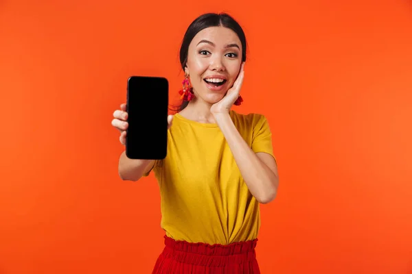 Izgatottan boldog fiatal nő pózol elszigetelt narancssárga fal háttérben mutatja megjelenítését mobiltelefon. — Stock Fotó