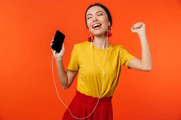 Piękna młoda kobieta pozowanie na białym tle na pomarańczowej ścianie tła za pomocą telefonu komórkowego słuchania muzyki. — Zdjęcie stockowe