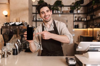 Kafe barda poz veren mutlu kahveci cep telefonunun sergilenmesini gösteren kapalı mekanda çalışıyor.