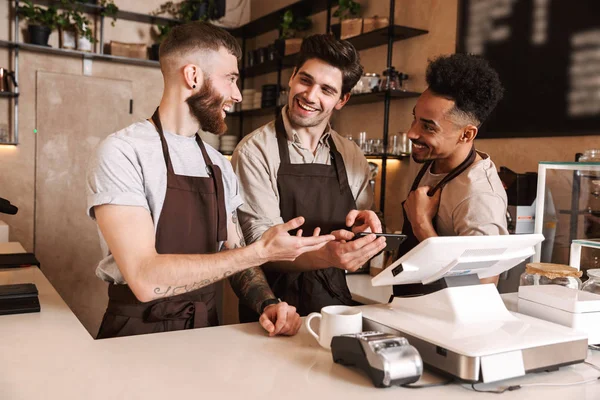 Three cheerful men baristas standing behind the counter — Stock Photo, Image