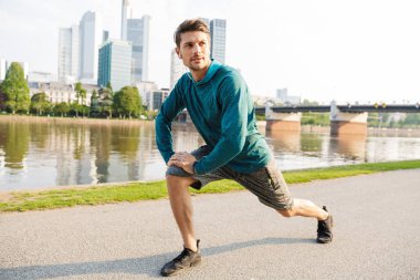 Çekici konsantre ciddi genç güçlü sporcu açık havada parkta kulaklıkile müzik dinlerken bacaklar için egzersizyapmak.