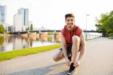 Şehir nehir kenarında çalışırken kulaklık kullanan ve ayakkabı bağcıklarını bağlayan mutlu adamın fotoğrafı