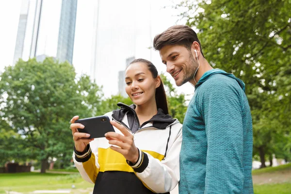 Foto av atletisk leende man och kvinna med hjälp av mobiltelefon medan du tränar i City Park — Stockfoto