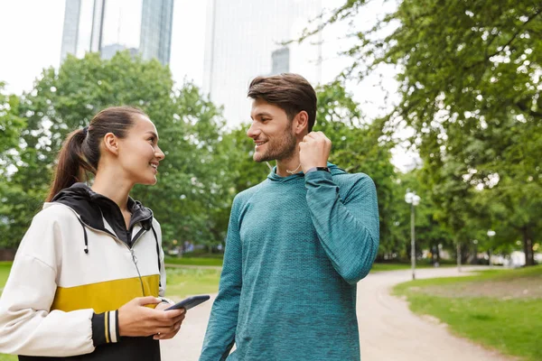 Фотографія спортивного усміхненого чоловіка та жінки, що використовує мобільний телефон та навушники під час розмови у міському парку — стокове фото