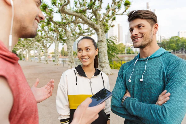Foto von sportlich lächelnden Menschen, die beim Training im Stadtpark mit dem Handy telefonieren und miteinander reden — Stockfoto