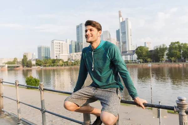 Foto de un joven feliz sonriendo y sentado en la barandilla mientras hace ejercicio cerca del paseo marítimo de la ciudad — Foto de Stock