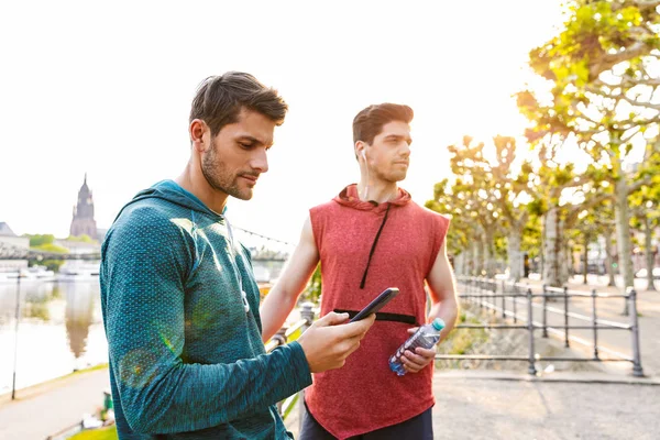 Foto de dos jóvenes enfocados en el uso de teléfonos celulares y auriculares mientras se ejercita en la ribera de la ciudad — Foto de Stock