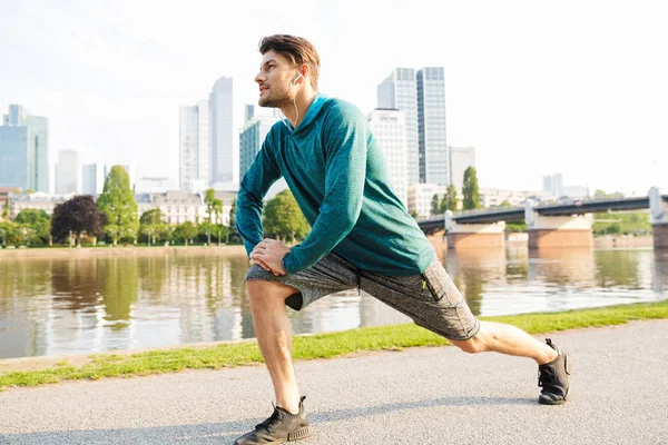 Привлекательный сконцентрированный серьезный молодой сильный спортсмен на открытом воздухе в парке слушает музыку с наушниками делают упражнения для ног . — стоковое фото