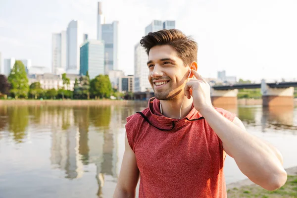 Photo of smiling athletic man using earphones while working out near city riverfront — Stock Photo, Image