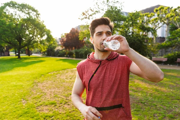 Zdjęcie kaukaski mężczyzna słuchania muzyki z słuchawek i wody pitnej podczas pracy w parku miejskim — Zdjęcie stockowe