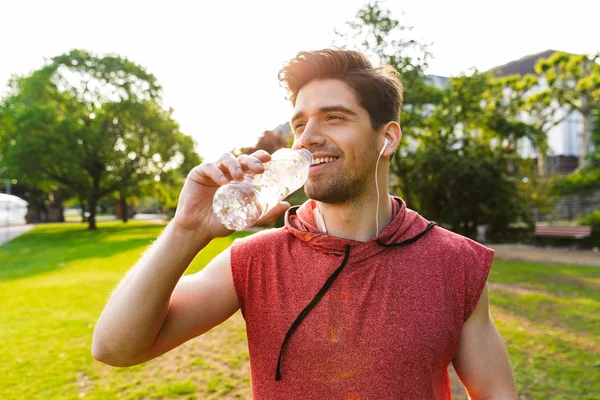 Foto eines fröhlichen Mannes, der Musik mit Kopfhörern hört und Wasser trinkt, während er im Stadtpark trainiert — Stockfoto