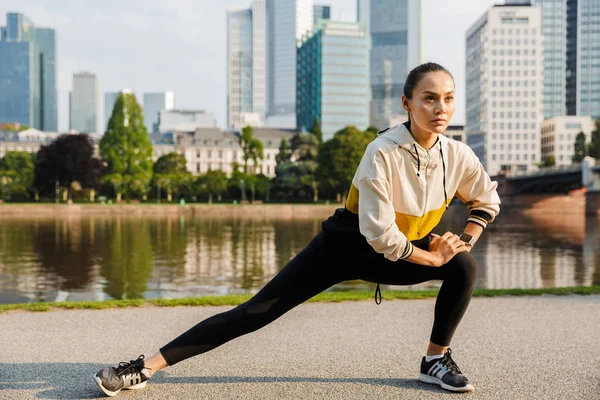 Foto de mujer deportiva concentrada estirando su pierna mientras hace ejercicio cerca de la ribera de la ciudad — Foto de Stock