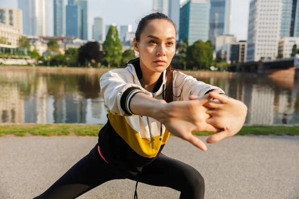 Foto de mujer atlética concentrada estirando sus piernas y brazos mientras se ejercita cerca de la ribera de la ciudad — Foto de Stock