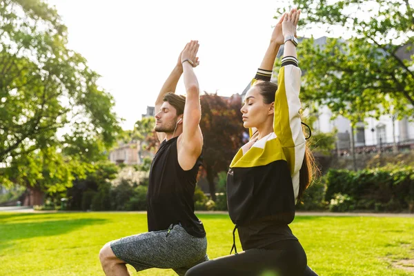 Foto de hombres y mujeres jóvenes enfocados haciendo ejercicios de yoga con los ojos cerrados mientras hacen ejercicio en el parque de la ciudad —  Fotos de Stock